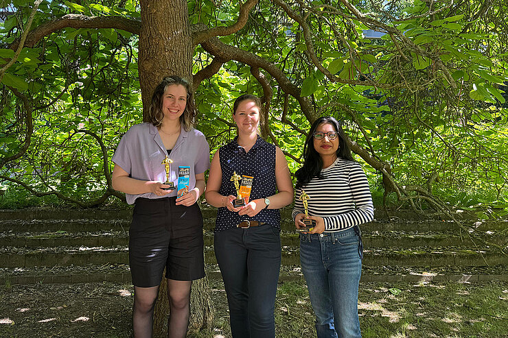 Three women stand under a tree, each holding a prize