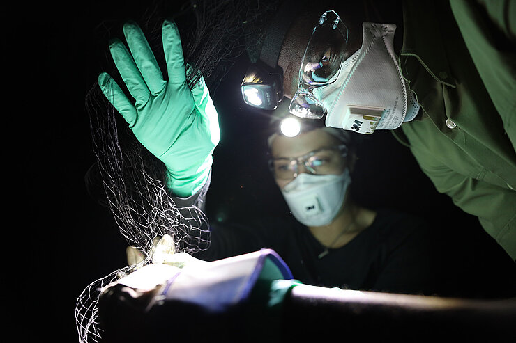 Two people wearing protective masks, goggles, and headlamps are examining a fine mesh material in the dark.