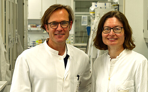 A man and a woman wearing white lab coats are smiling at the camera while standing in a laboratory
