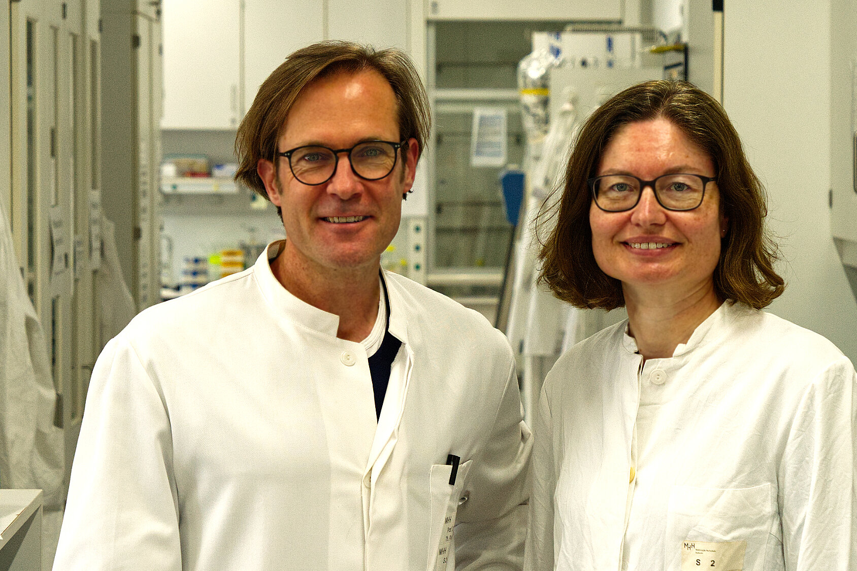 A man and a woman wearing white lab coats are smiling at the camera while standing in a laboratory