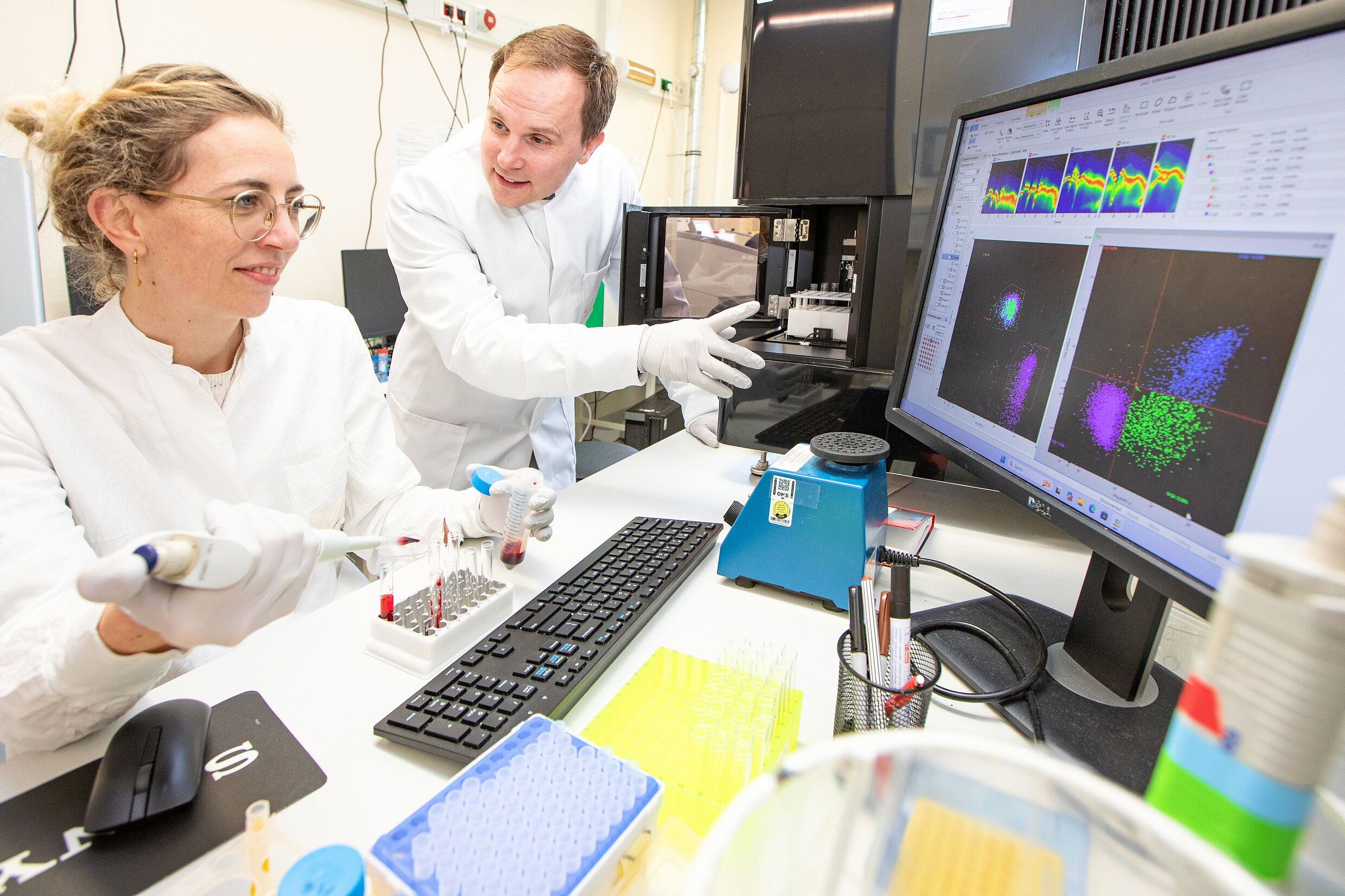 A man and a woman are both wearing lab coats. They are seen in a laboratory, looking at a screen
