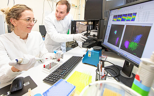 Ein Mann und eine Frau haben je einen Kittel an. Sie sind in einem Labor zu sehen an einem Bildschirm. 